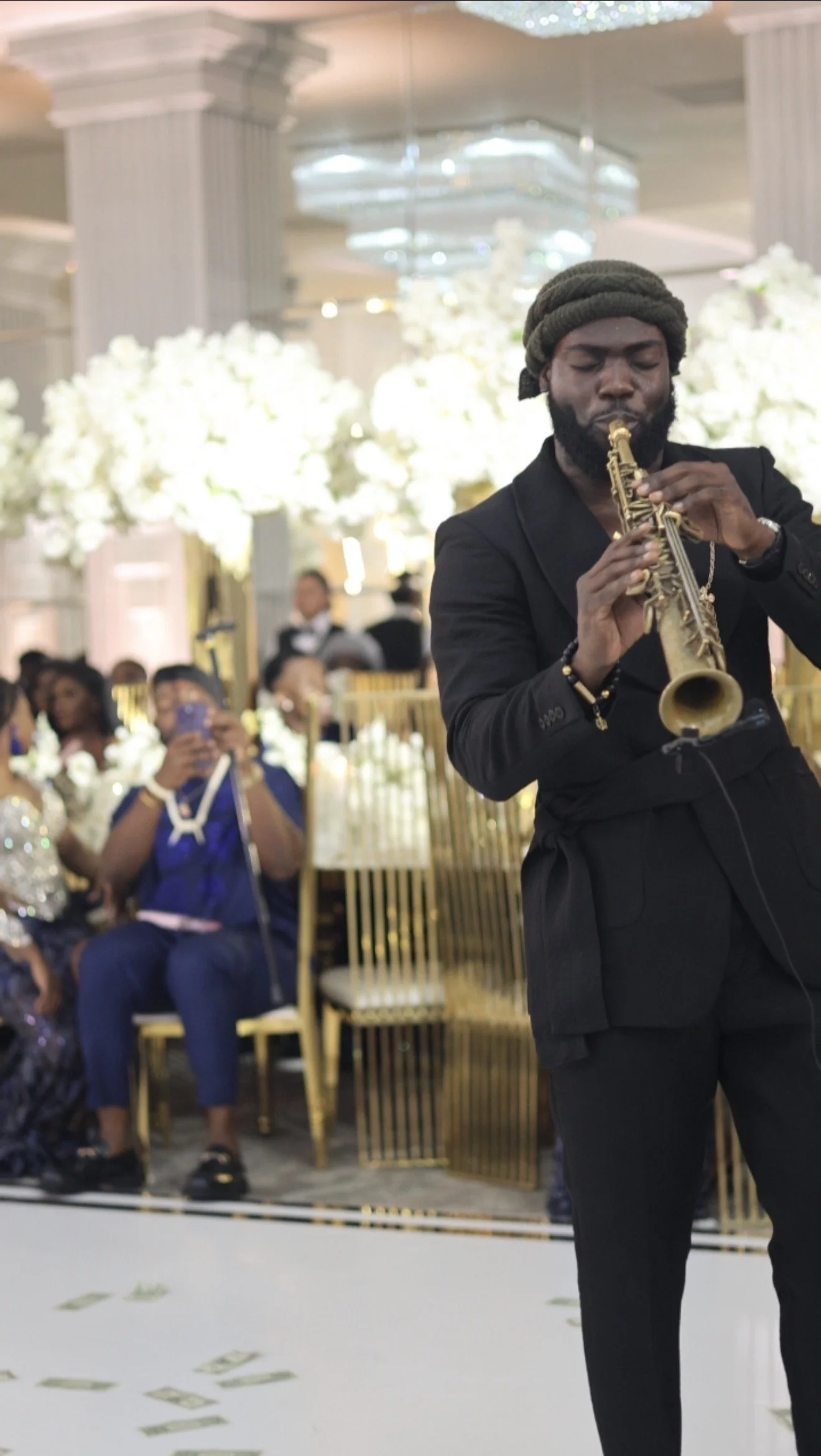 A man in black suit playing saxophone at an event with guests seated in the background.