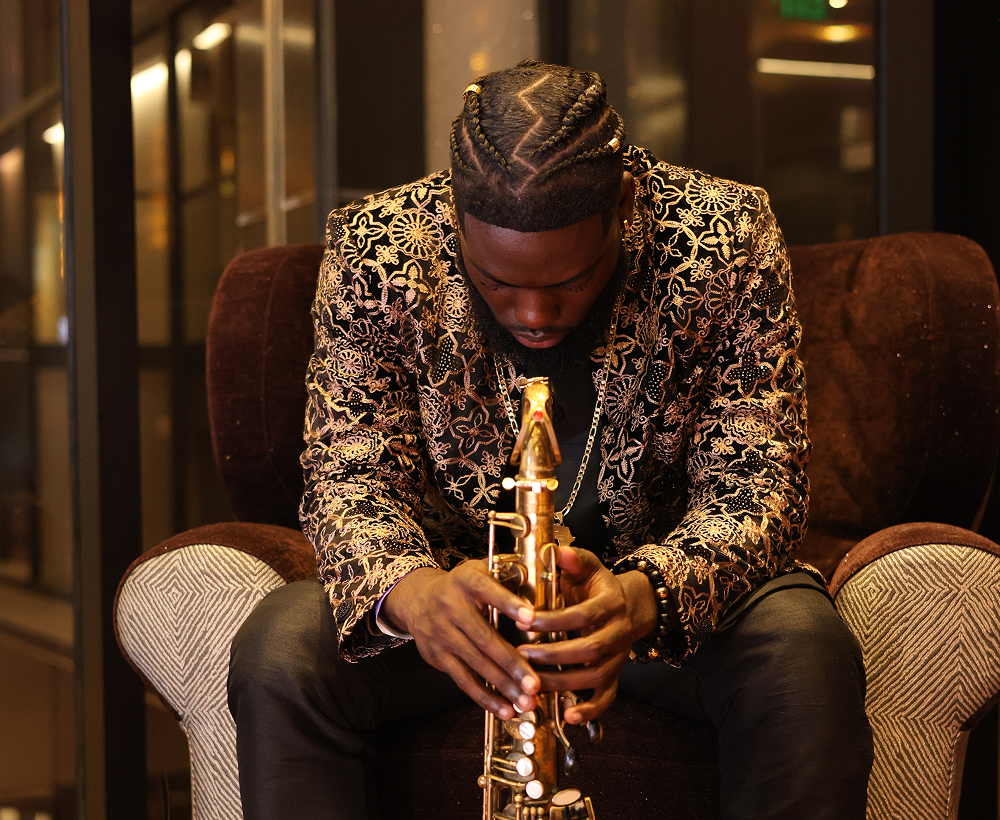 A man with braided hair wearing a floral patterned jacket sitting on a dark brown chair, holding a saxophone in a dimly lit room.