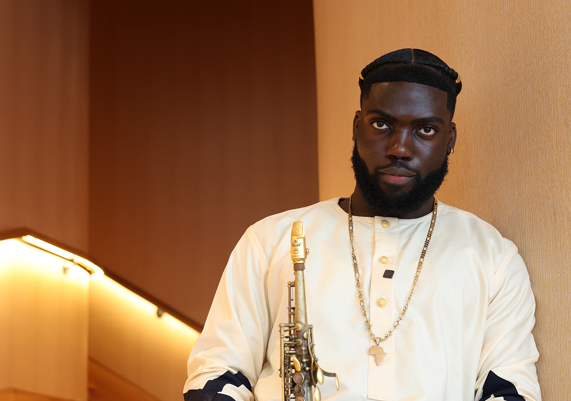 A man with dark skin, black braided hair, and a beard, holding a saxophone, sitting against a wooden wall with warm lighting.