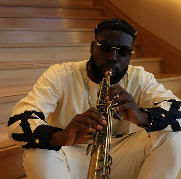 Person sitting on wooden stairs playing a saxophone, wearing sunglasses, a cream-colored outfit with black accents, and a gold chain.