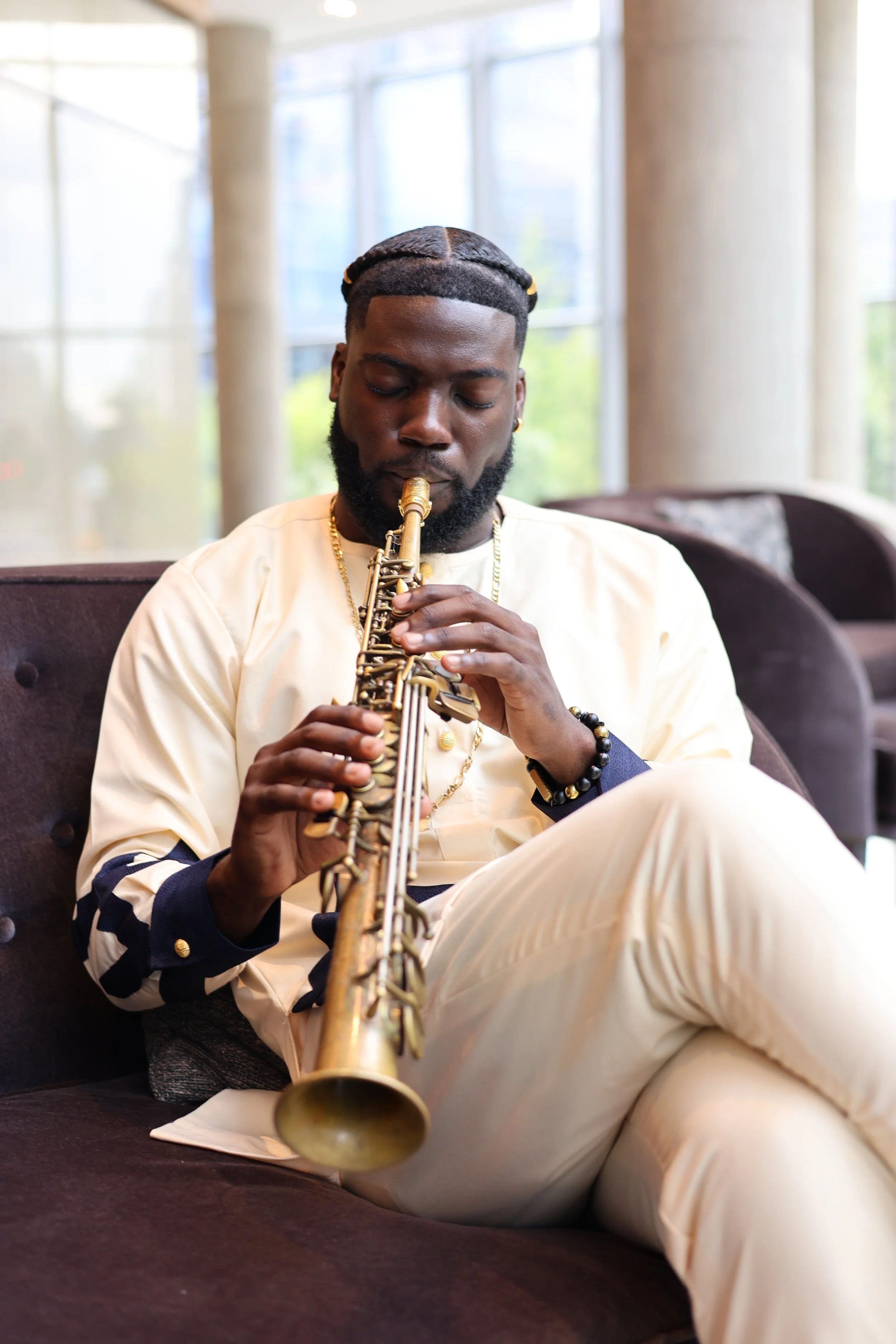 A man with styled hair and beard, wearing a cream-colored shirt with black and gold embellishments, is sitting on a couch playing a gold saxophone in a bright room with large windows.