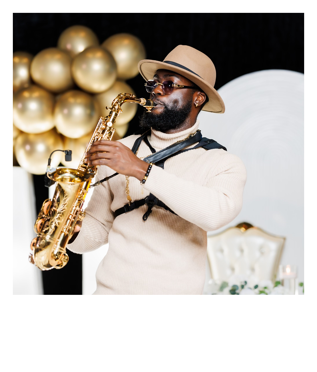 A man wearing a tan hat, sunglasses, and a beige sweater playing a gold saxophone indoors with decorative gold balloons and a white ornate backdrop.