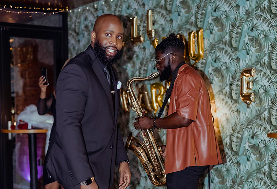 Two men at a celebration, one in a suit and the other playing a saxophone, with balloons spelling 'LOVE' in the background.