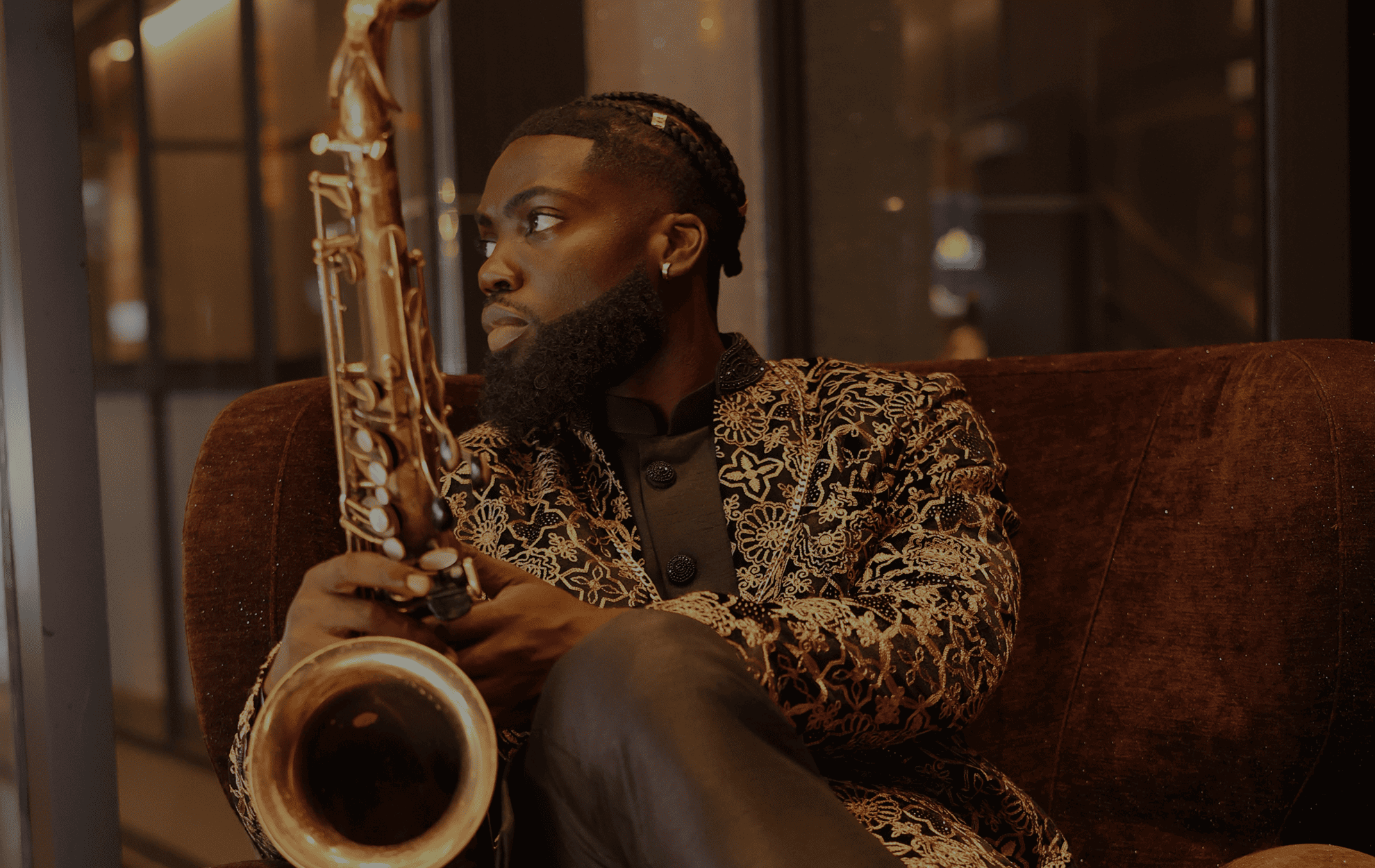 A man with braided hair, a full beard, and earrings, sitting on a brown velvet sofa, holding a musical instrument, possibly a saxophone, in a warmly lit room with glass-paneled walls.