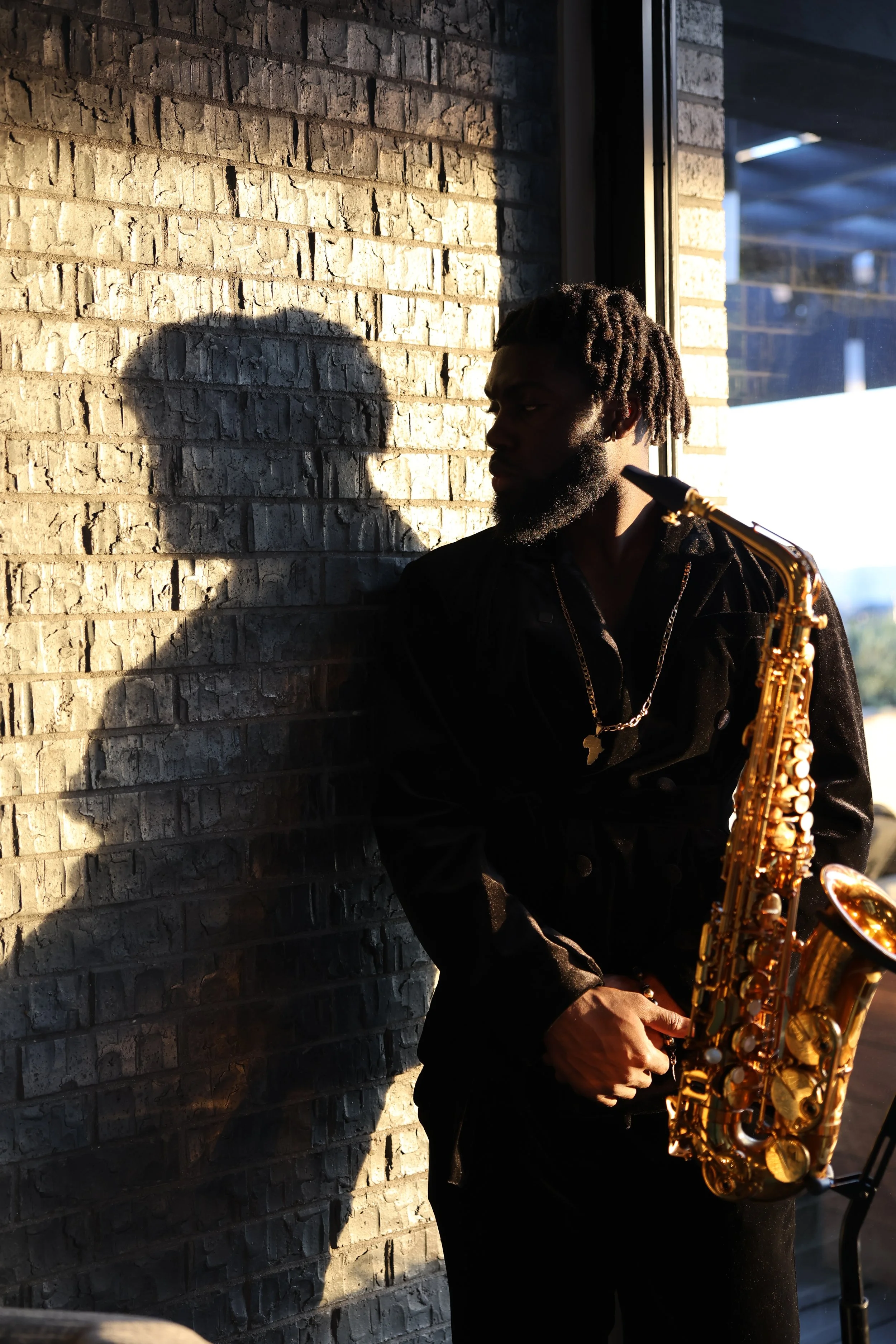 A man with dreadlocks and a beard standing near a brick wall in sunlight, holding a gold saxophone, wearing a black jacket and gold chain, shadows cast on the wall behind him.
