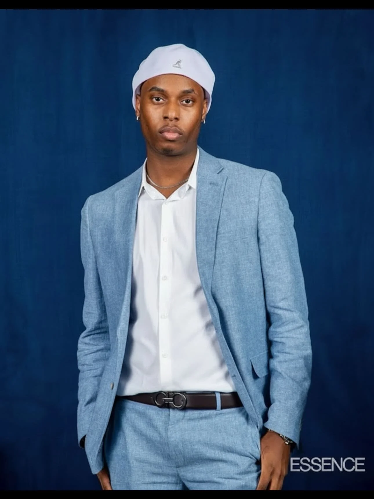 A person wearing a light blue suit and white shirt with a white beanie, standing against a dark blue background with the word 'ESSENCE' at the bottom right.