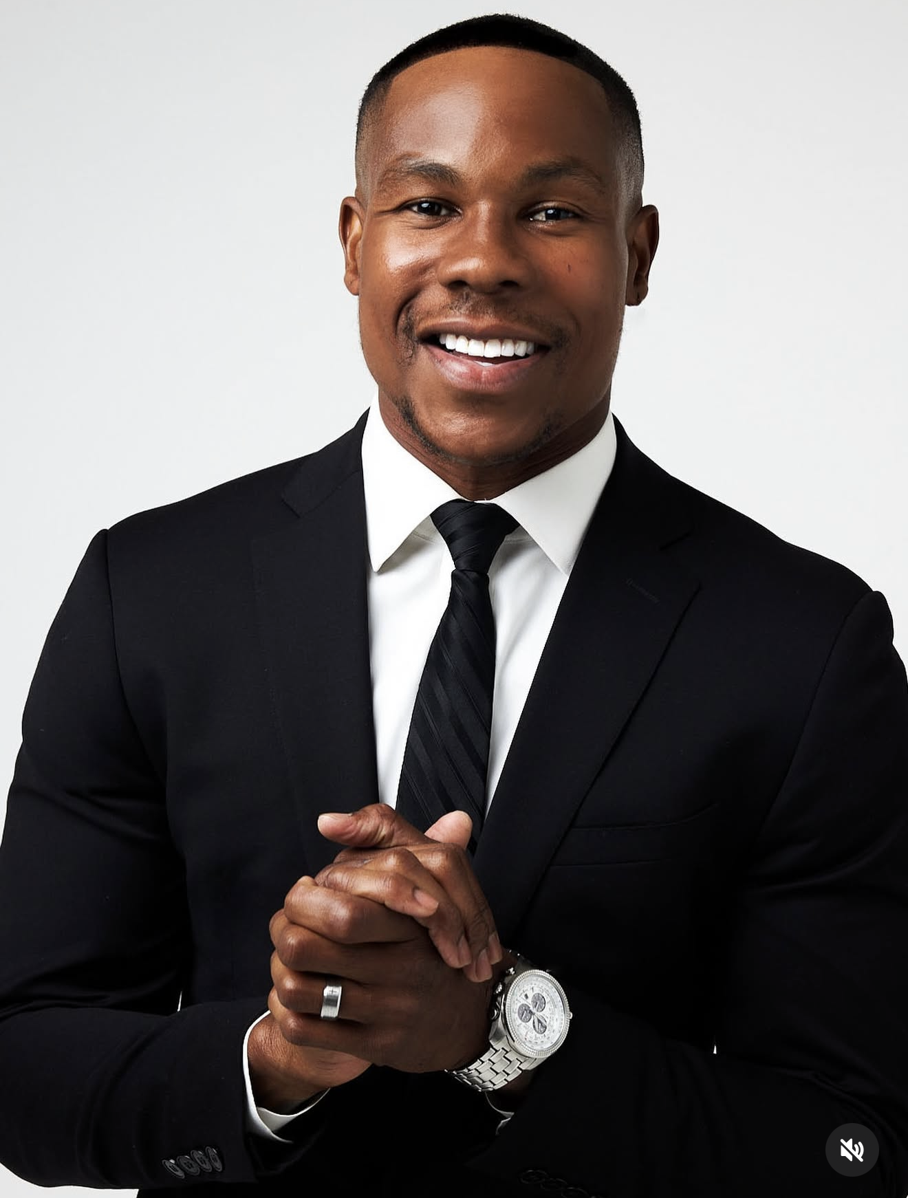 A confident African American man in a black suit, white shirt, and black striped tie, smiling and clasping his hands, wearing a silver wristwatch and ring, in front of a plain gray background.
