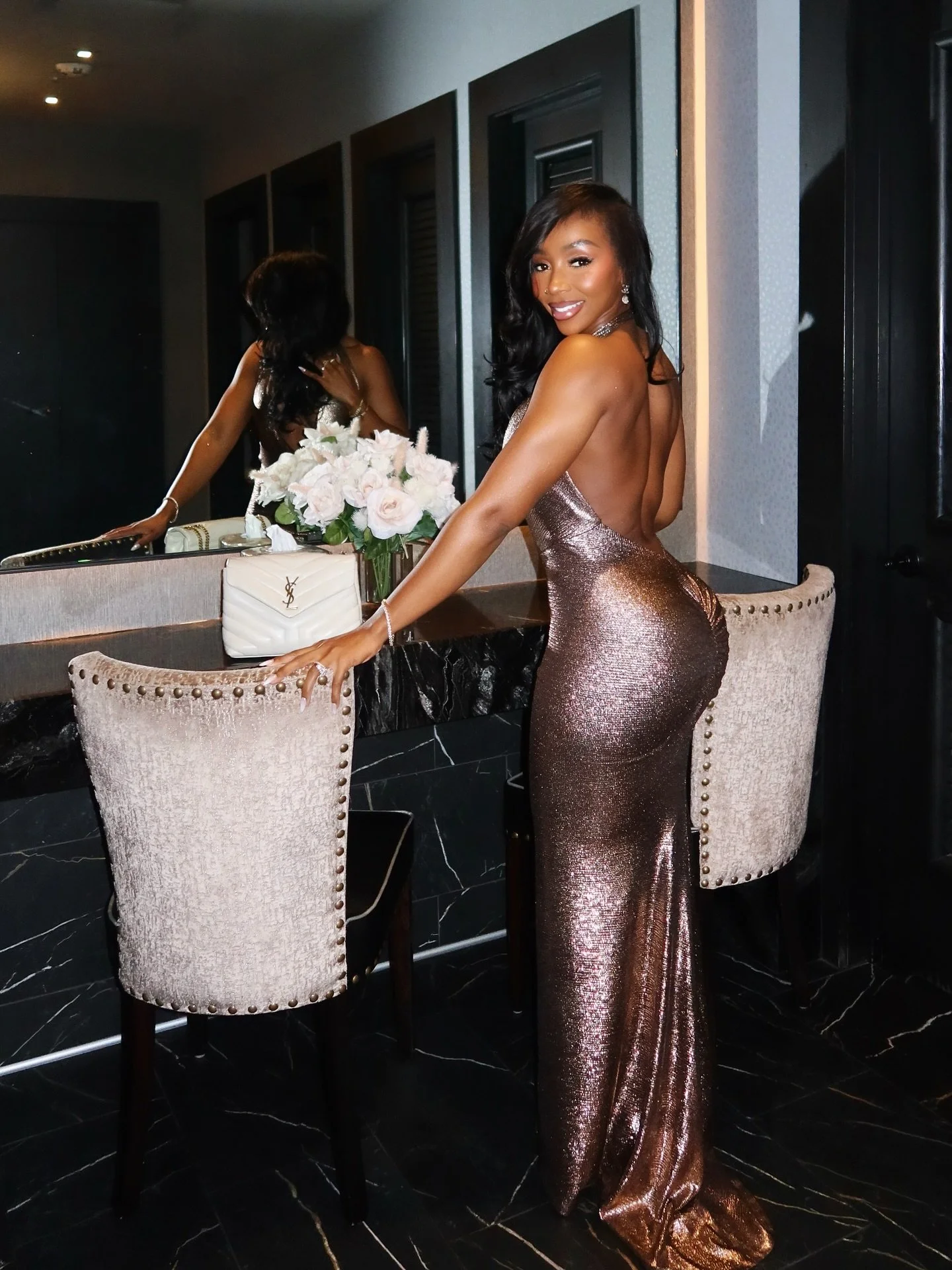 A woman in a shimmering, backless, bronze evening gown standing in front of a mirror, smiling, with a bouquet of roses and a white designer purse on a vanity table.