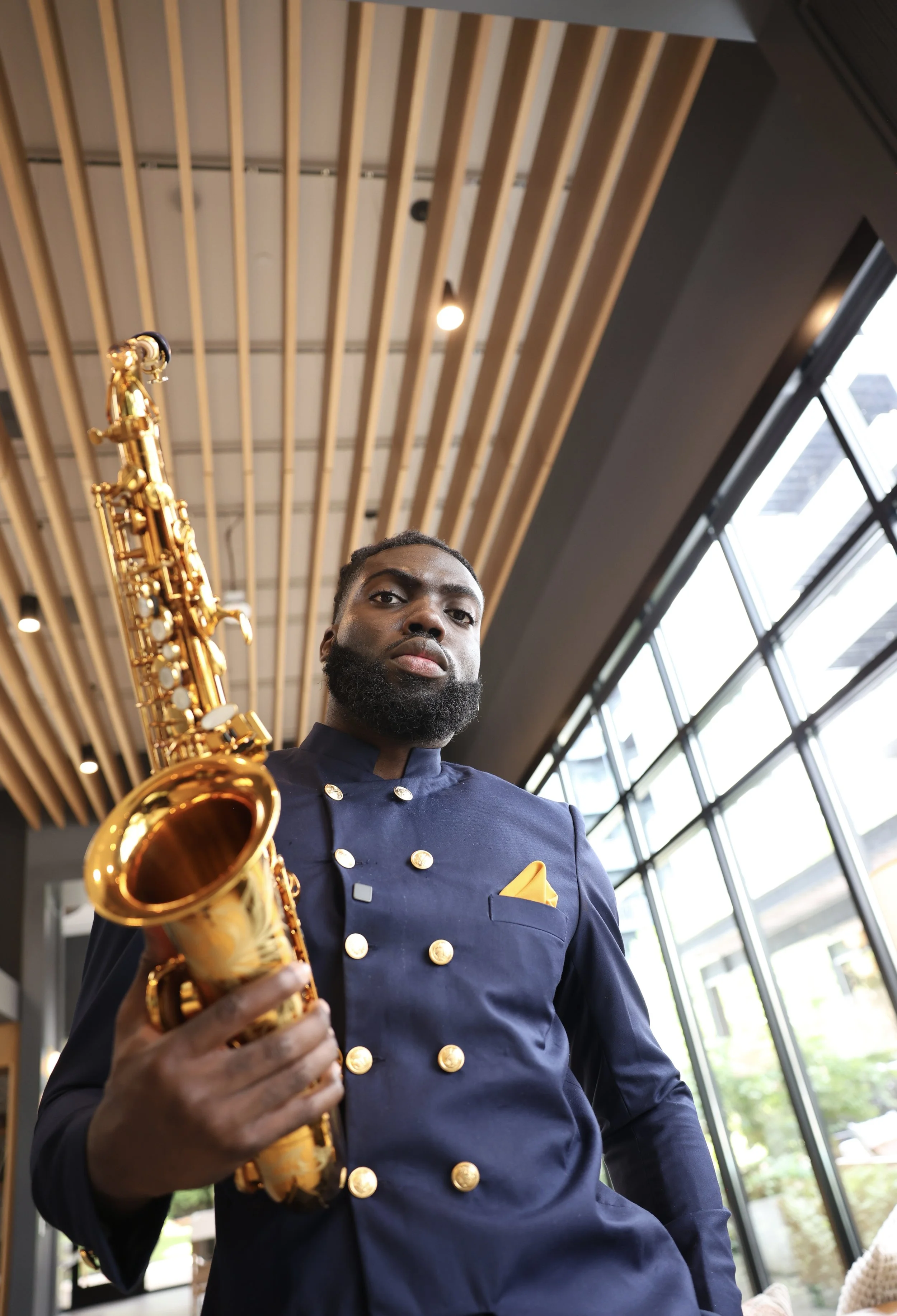 A man in a navy blue chef's uniform holding a gold-colored saxophone inside a modern building with large windows and wooden ceiling slats.