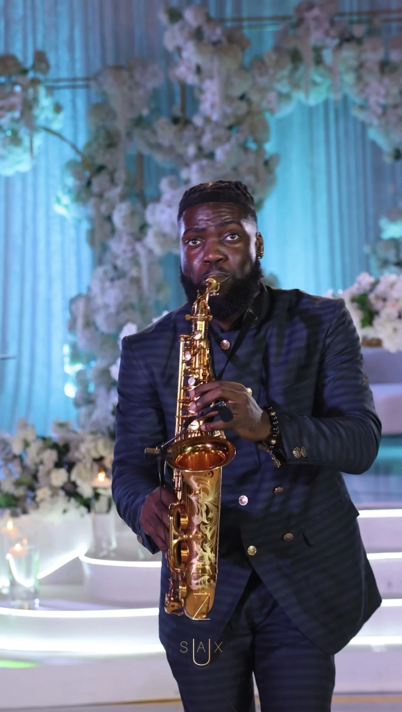 Man playing a gold saxophone at a decorated event with white flowers and blue background.