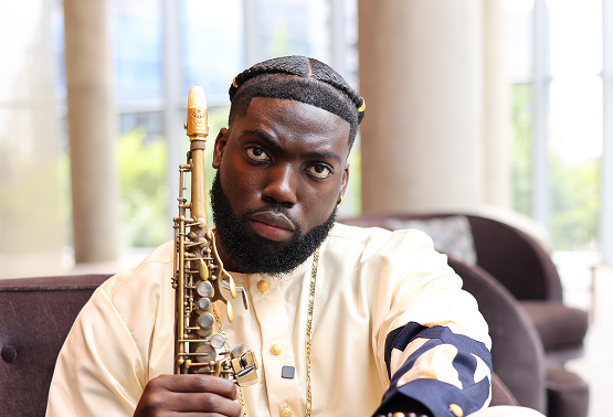 A man with braided hair and a beard sitting on a sofa holding a saxophone, indoors with large windows in the background.