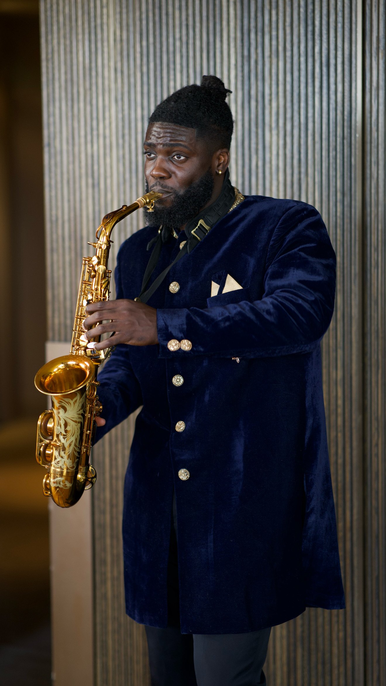 A man wearing a dark blue velvet blazer with gold buttons and a black shirt, playing a gold saxophone.