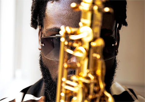 Close-up of a man with sunglasses and a beard, playing a shiny gold saxophone.