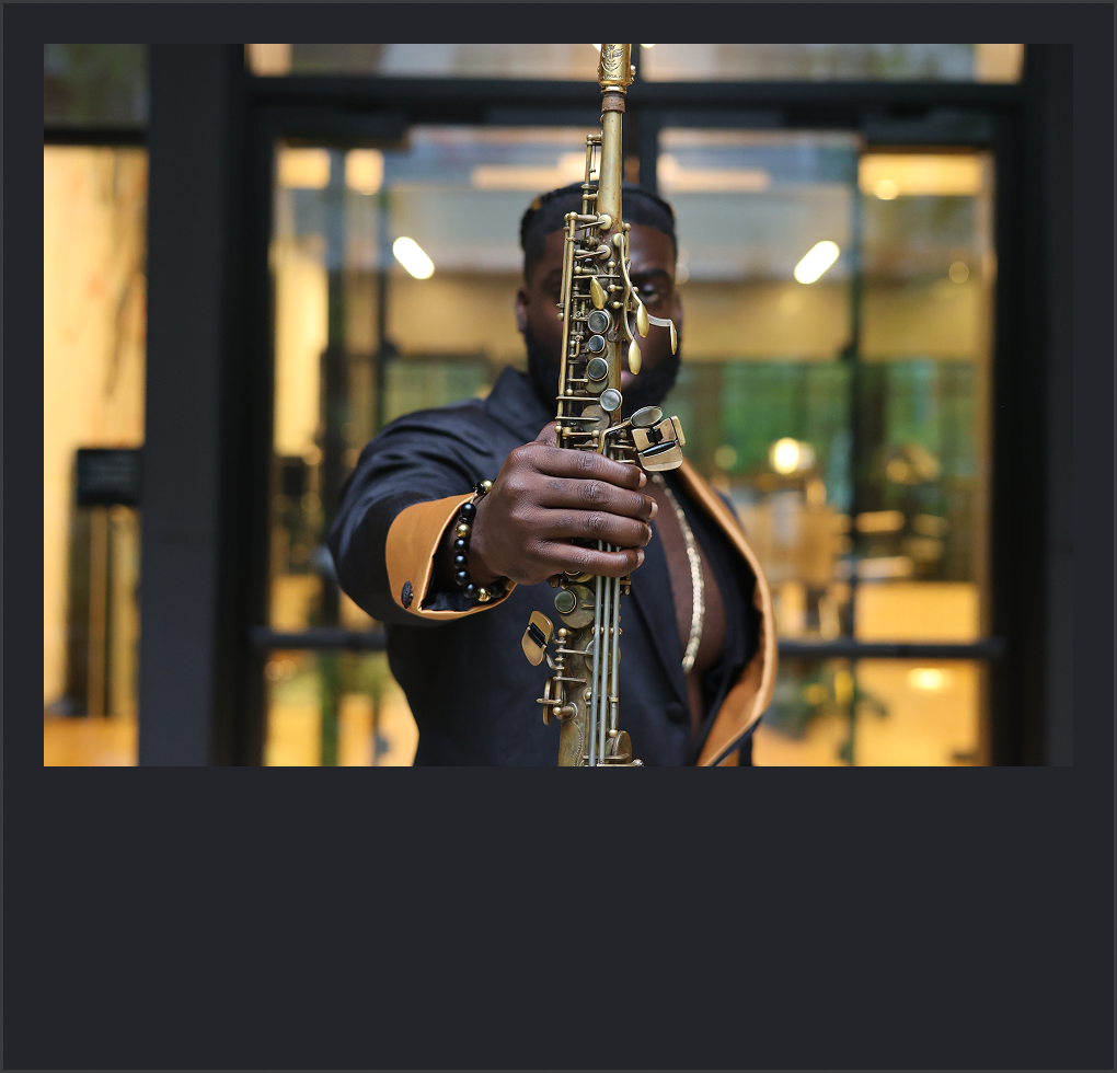 Man holding a gold-colored saxophone in front of him, partially hiding his face, indoors with a glass door in the background.