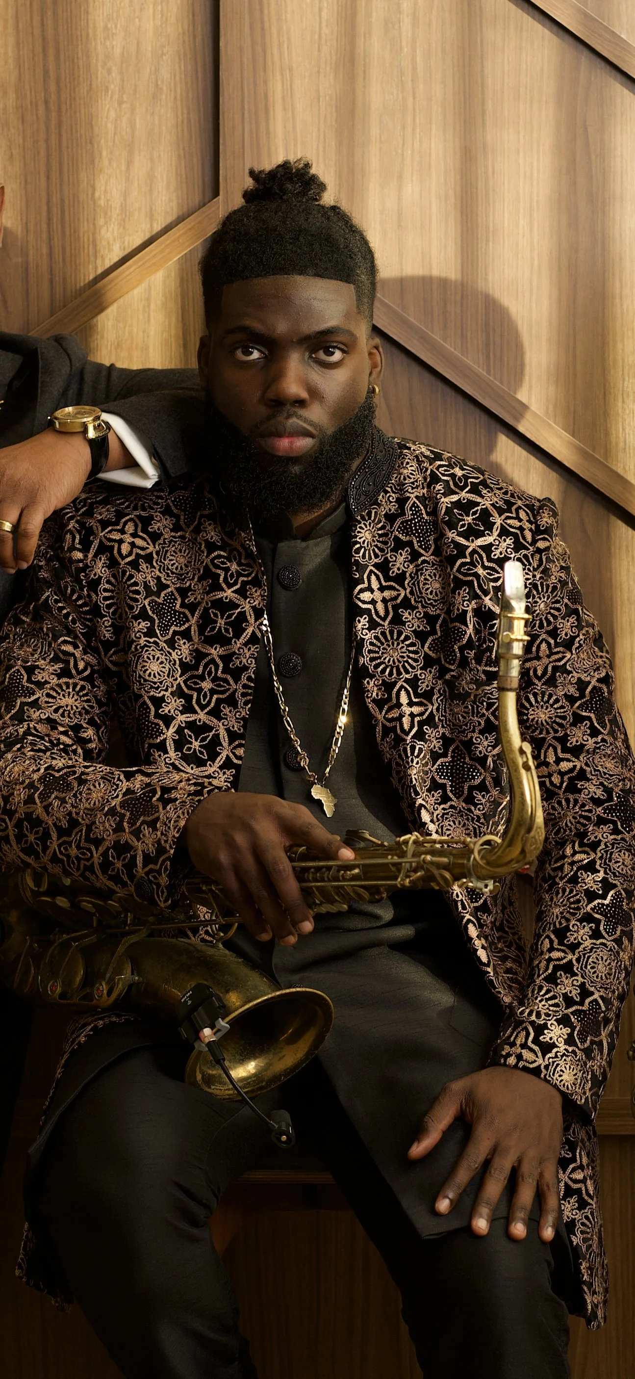 Young man with dark skin, wearing a patterned blazer, black shirt, and gold jewelry, holding a saxophone while sitting against a wooden wall.
