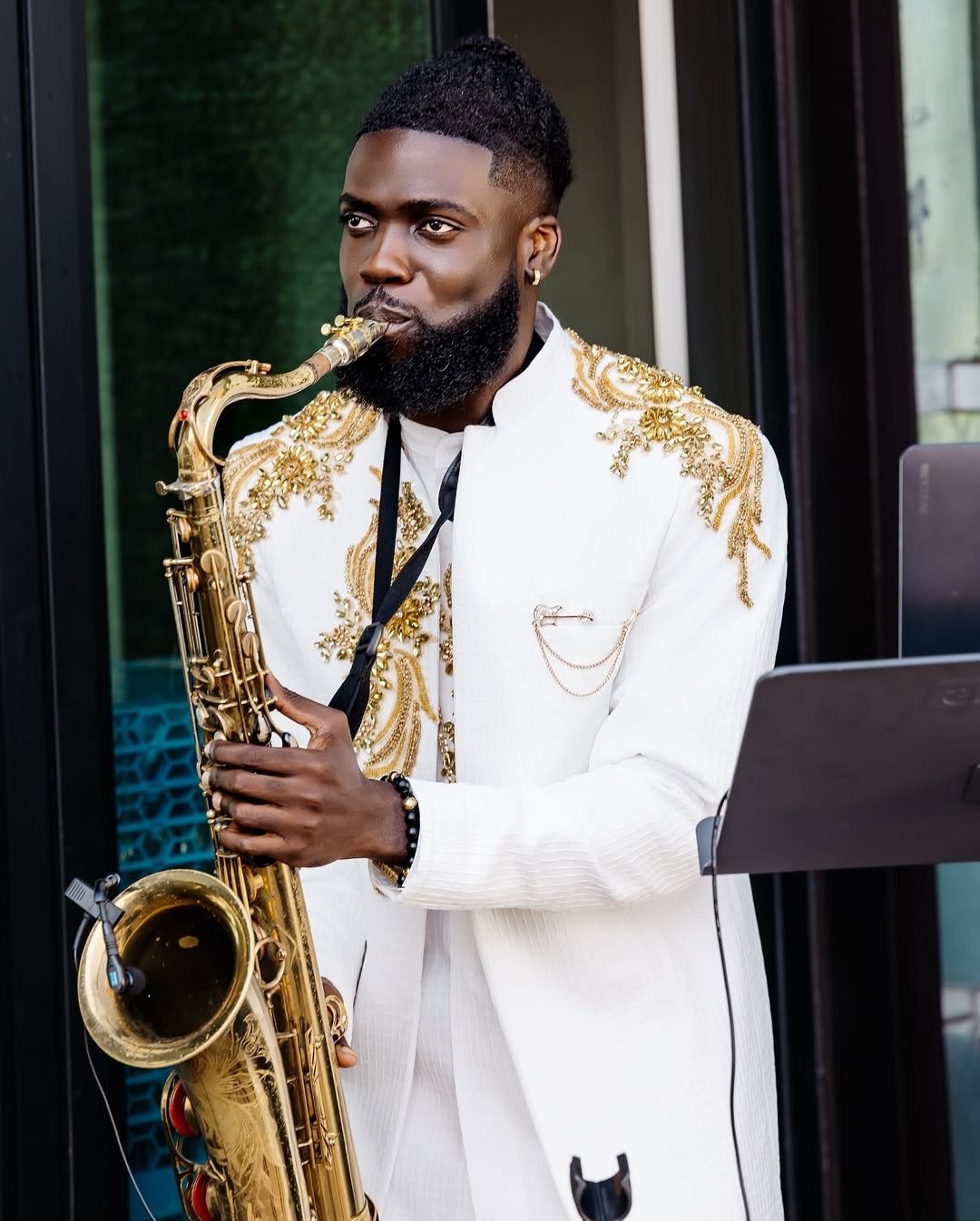 A man with a short, curly hairstyle and a beard, dressed in a white suit with gold embroidery, playing a saxophone.