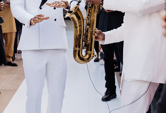 Two people are performing a musical act with one holding a saxophone and the other gesturing with their hands, both dressed in white suits, in a crowd setting.
