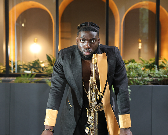 A man in a black suit with gold accents, holding a saxophone, stands in front of garden beds with buildings and arches in the background.
