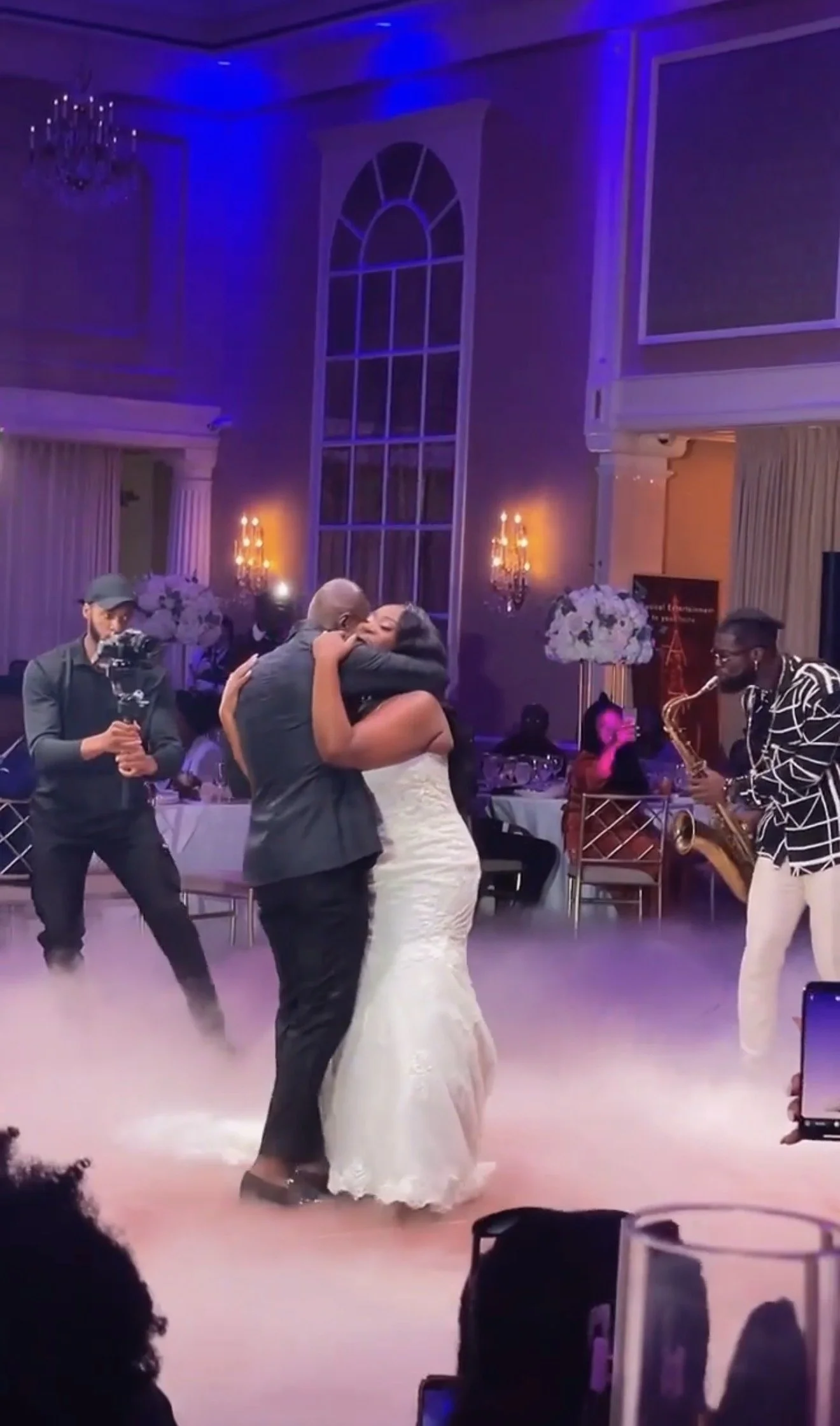 Couple dancing closely at wedding reception with a musician playing the saxophone nearby, in an elegant decorated venue with large window and floral arrangements.