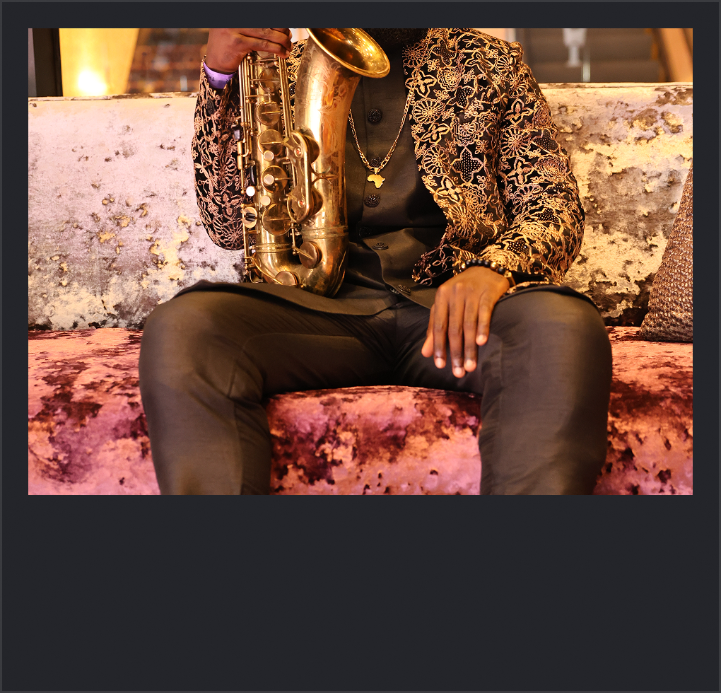 Person sitting on a patterned velvet couch holding a saxophone, dressed in a floral blazer, black shirt, and dark pants, with jewelry visible.