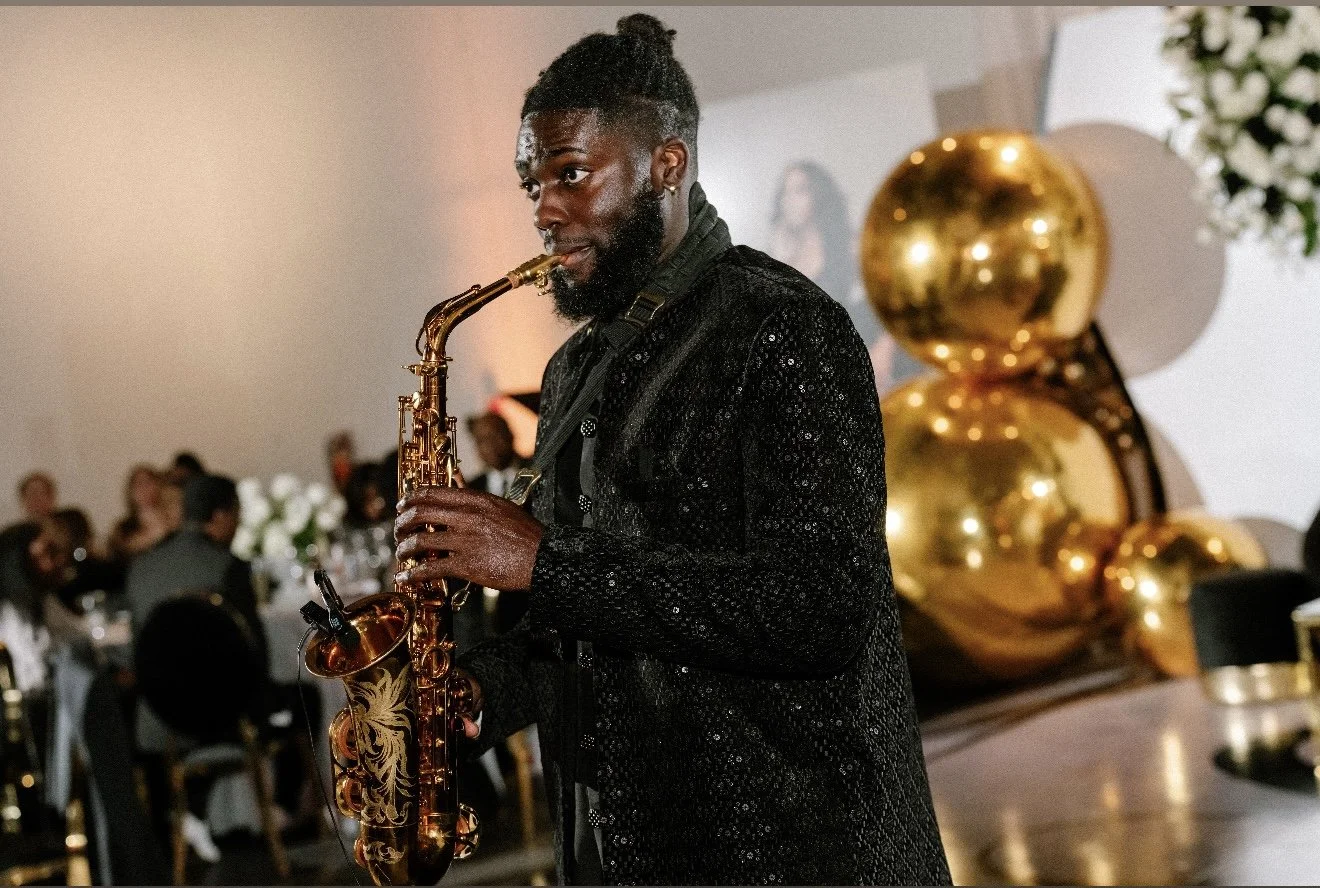 A man playing a saxophone at an indoor event, with gold balloons and floral decorations in the background.