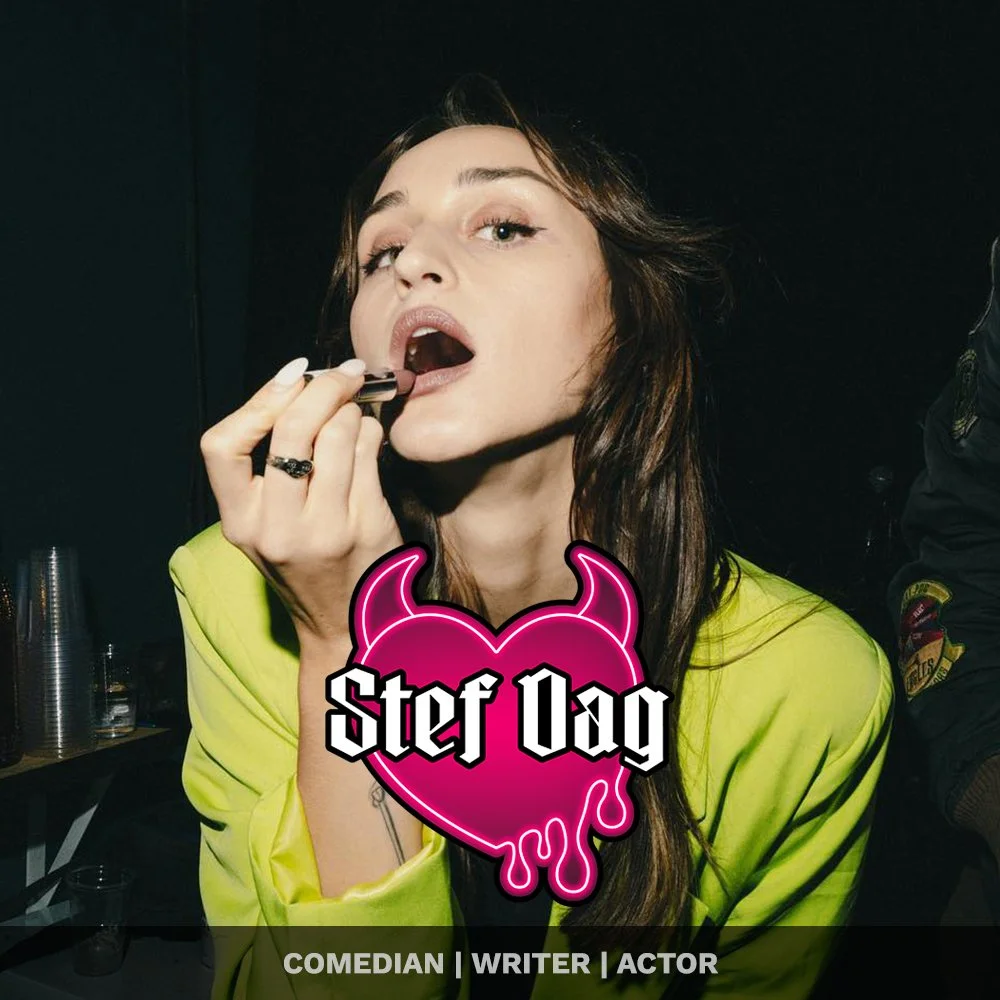 A woman with long brown hair applying lipstick in front of a mirror, wearing a yellow jacket, with a neon pink heart with devil horns and the words 'Stef Day' on her chest, against a dark background.