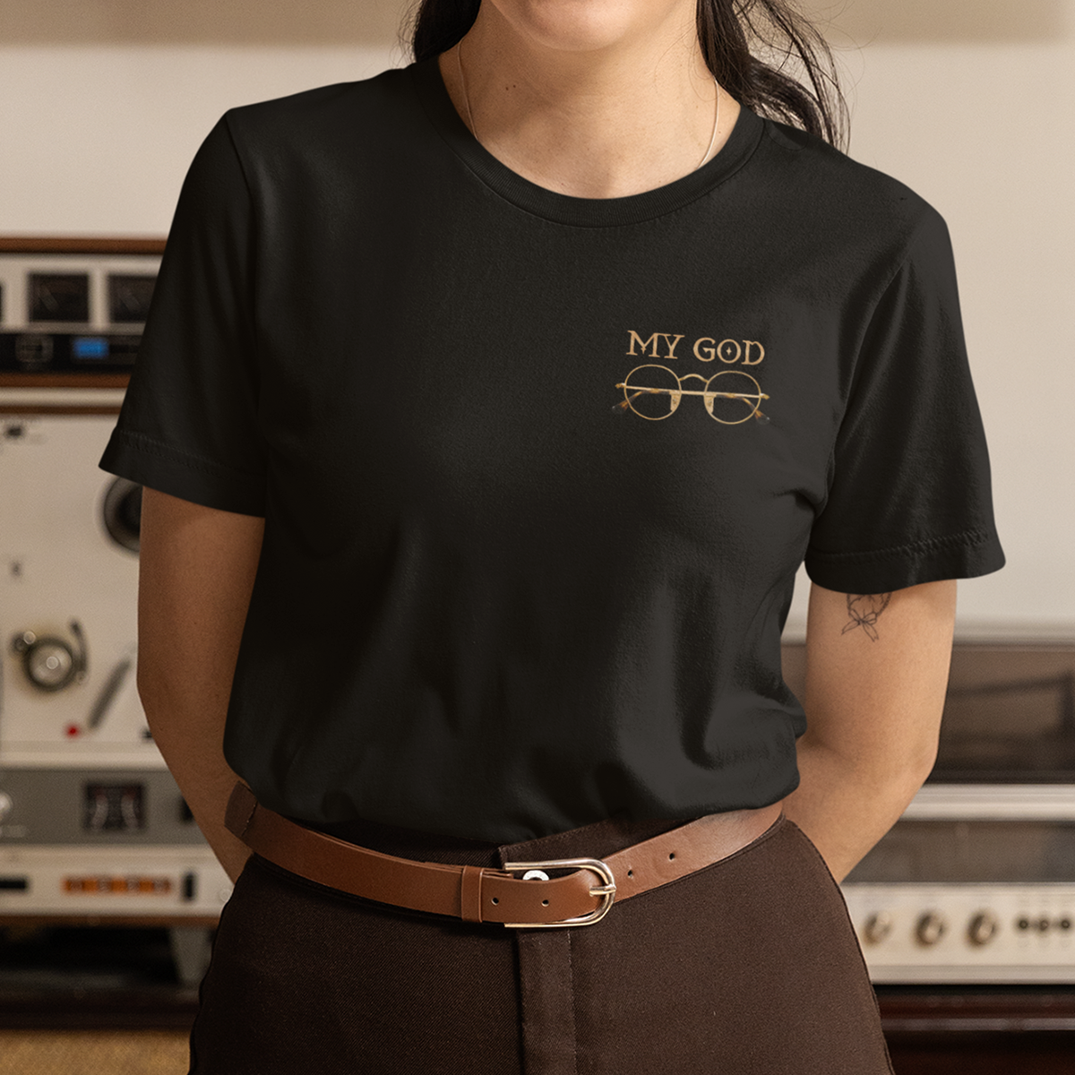 A woman wearing a black T-shirt with the words "MY GOD" and an illustration of glasses on it, standing in front of vintage audio equipment.