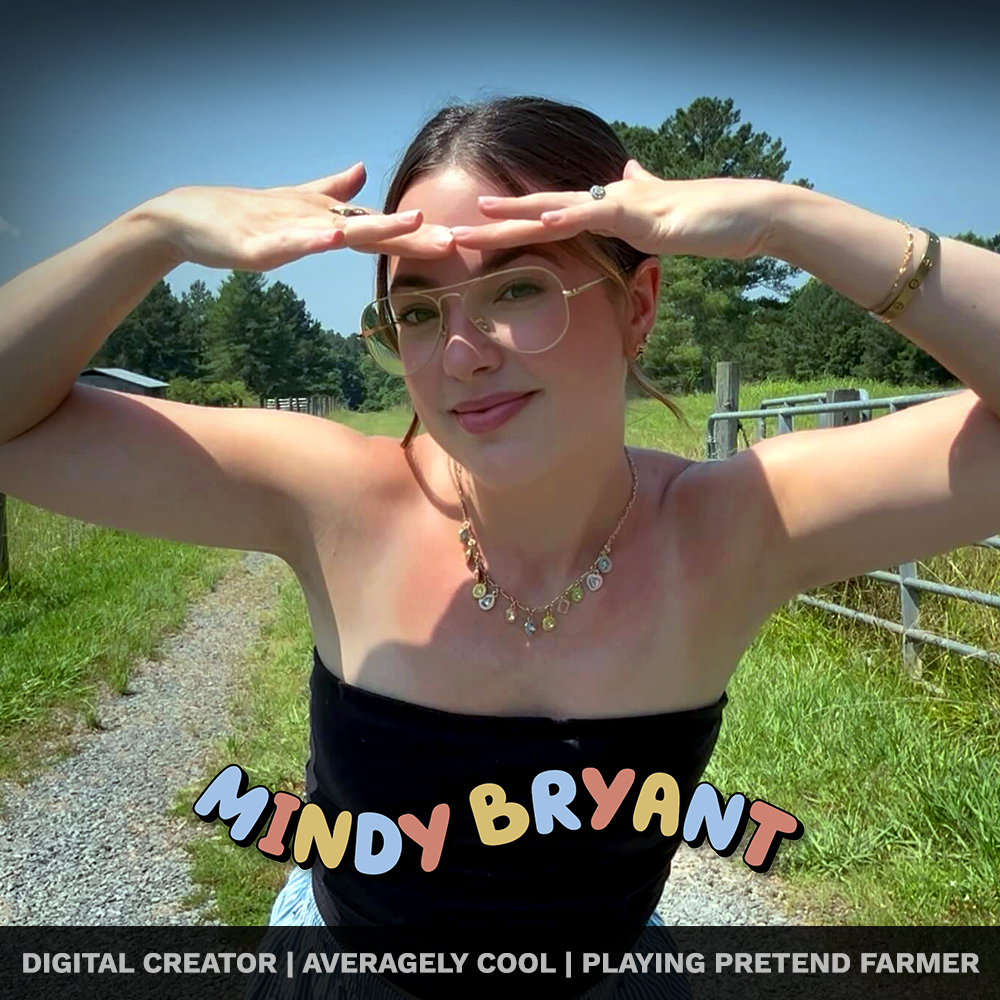 A woman wearing glasses and jewelry, smiling while standing outdoors on a sunny day, with greenery and a fence in the background, holding her hands above her eyes as if shielding from the sun.