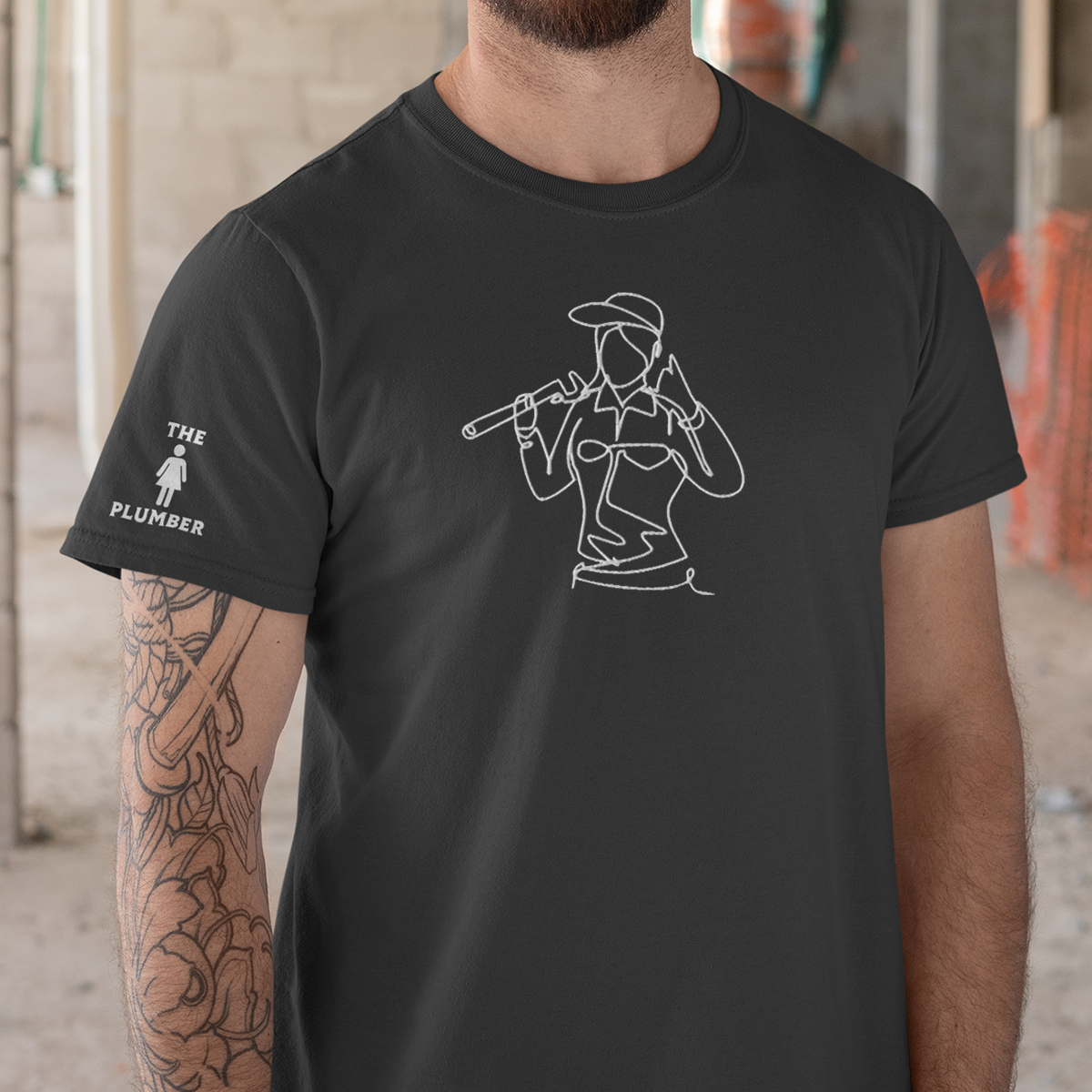 Close-up of a man wearing a black T-shirt with a white line drawing of a woman holding a pipe wrench over her shoulder, wearing a cap and sunglasses. The T-shirt has text on the sleeve that reads "THE PLUMBER" with a small icon of a woman.