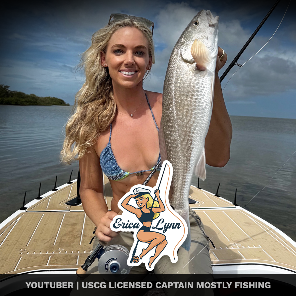 A woman with long blonde hair, wearing a blue bikini top, holding a large fish on a boat in a body of water during the day. She is smiling and standing on a wooden boat deck, with a cloudy sky in the background. There is a sticker of a cartoon woman 