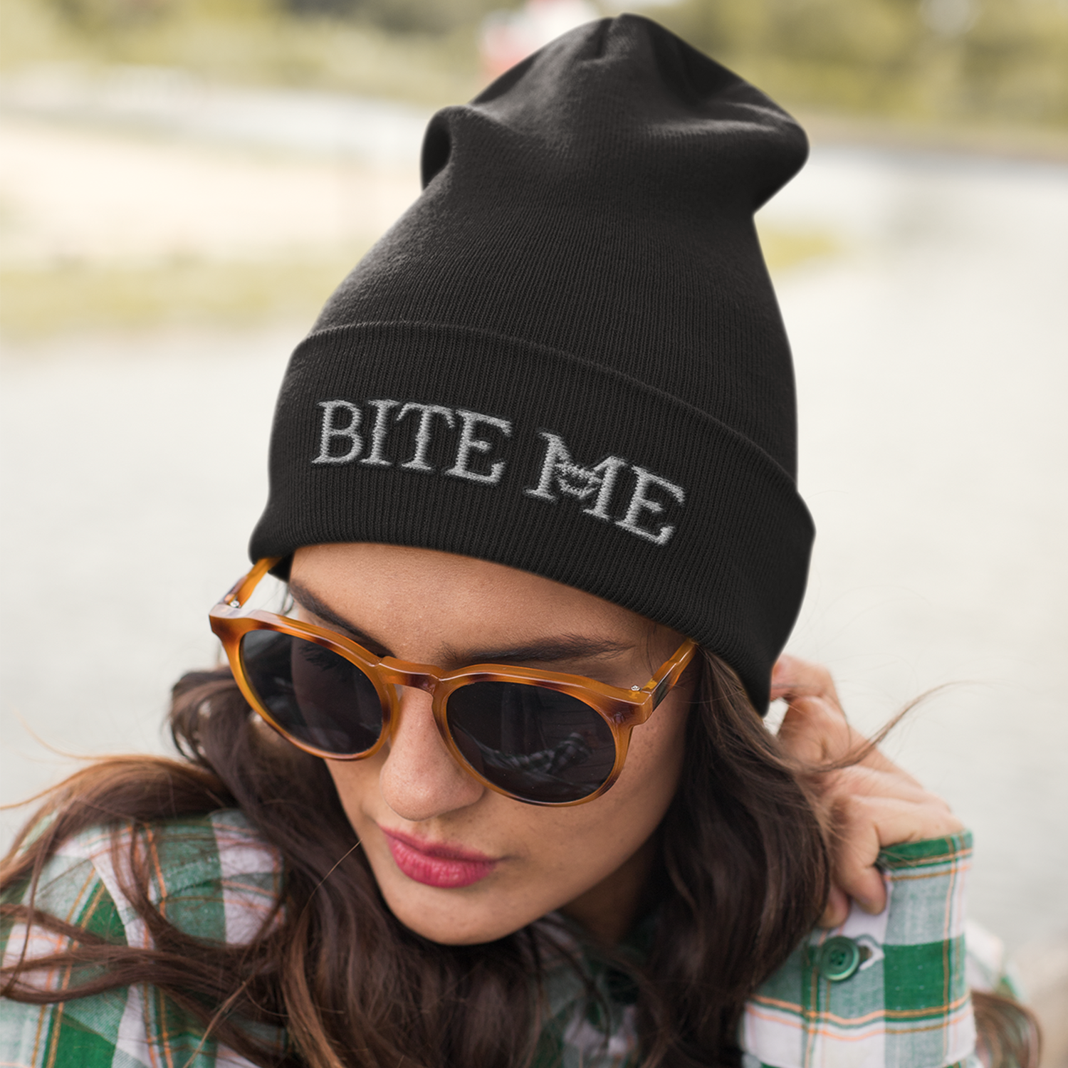 A woman with long brown hair wearing a black beanie with the words "BITE ME" embroidered on it, orange sunglasses, and a green plaid shirt, outdoors near a body of water.