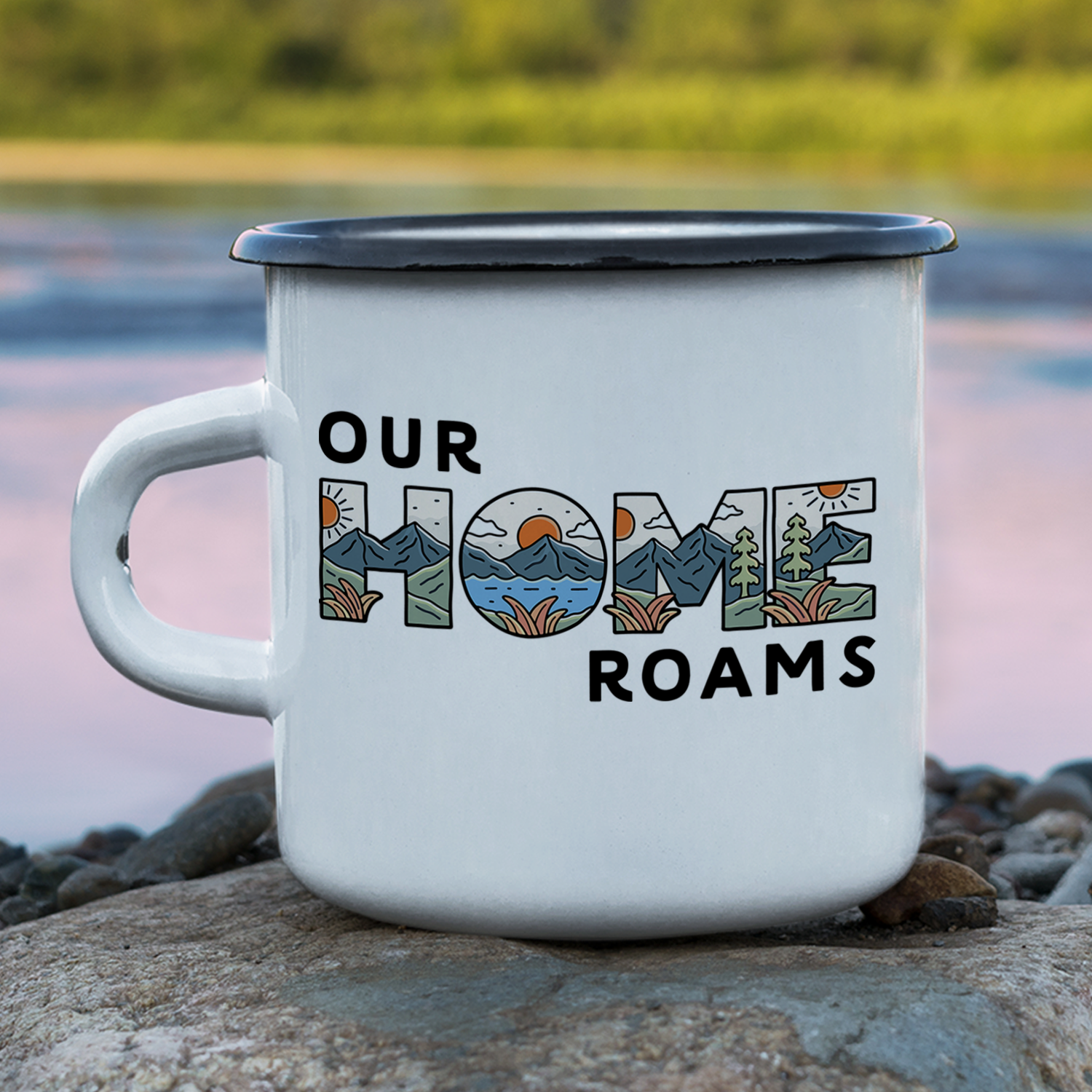 A white enamel mug with black rim on a rocky surface near water, displaying 'Our Home Roams' with scenic mountain, lake, and forest illustrations inside the text.