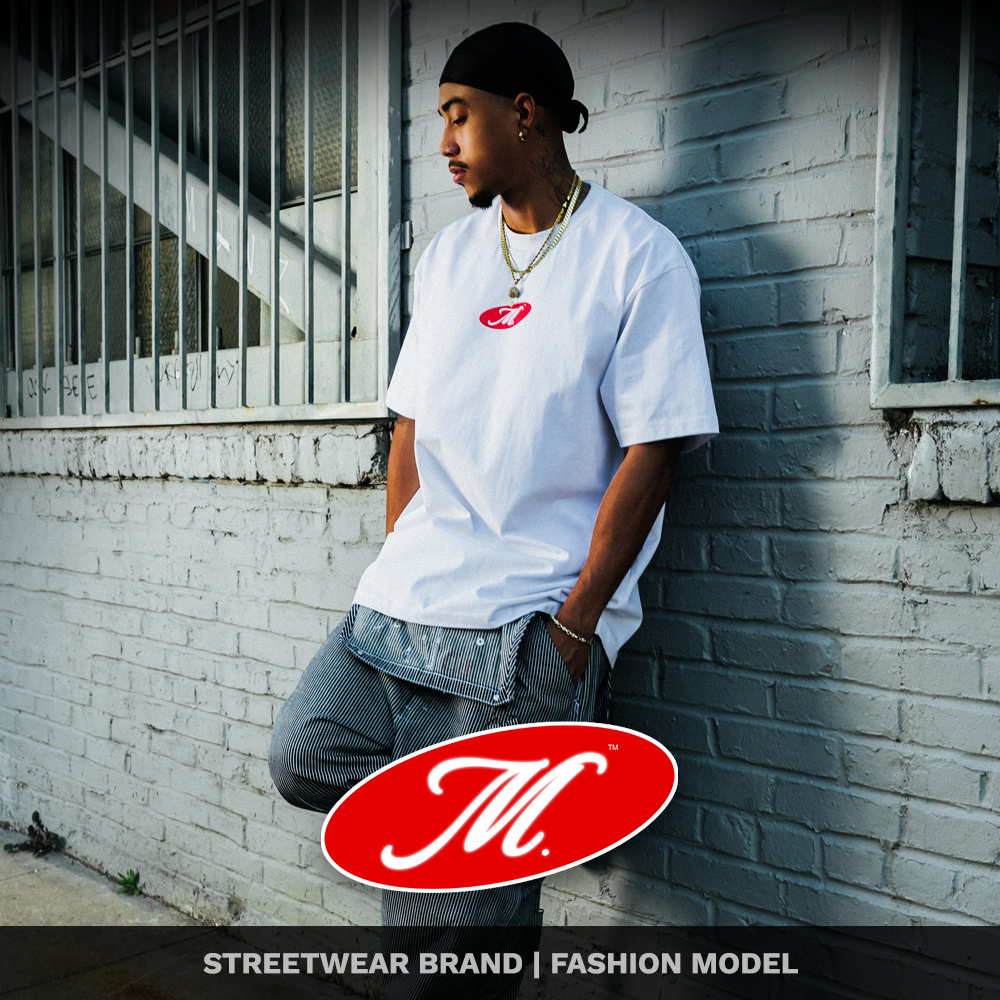 A young man with a tattooed neck, wearing a white T-shirt with a red 'M' logo, multiple gold chains, and a black headband, leaning against a white brick wall near a window with metal bars.