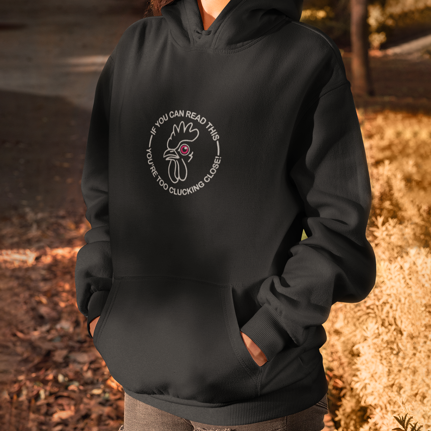Person wearing a black hoodie with a circular graphic of a cartoon chicken and the text 'If you can read this, you're too clucking close!' printed on front, standing outdoors at dusk with trees and fallen leaves in the background.