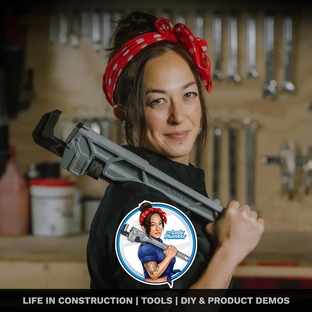 A woman with a red polka dot headband holding an oversized adjustable wrench over her shoulder, standing in a workshop with tools hanging on a wall behind her.