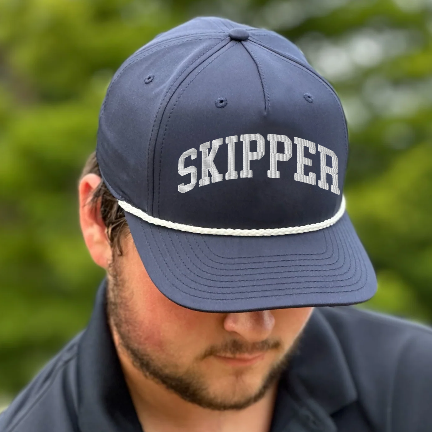 A person wearing a navy blue cap with the word 'SKIPPER' embroidered on it, outdoors with blurred green foliage in the background.