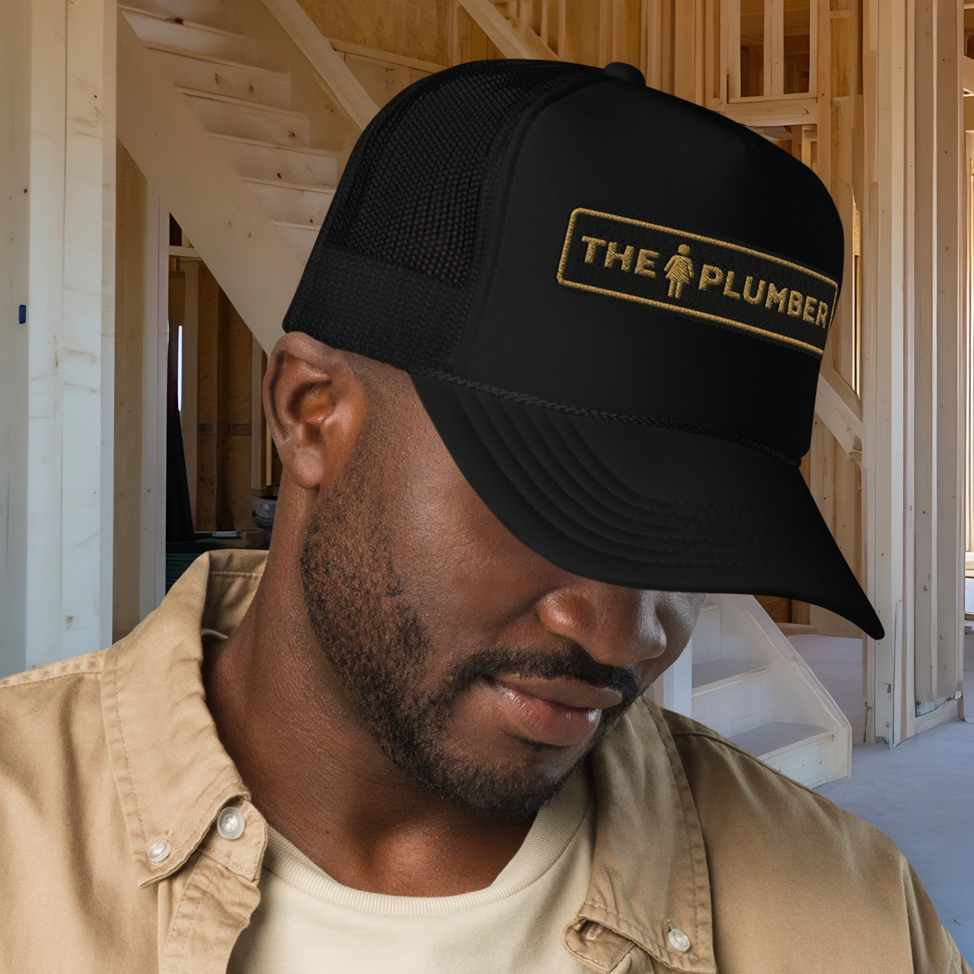 A man wearing a beige work shirt and cap with a patch that reads 'The Plumber' in a construction site or unfinished building.