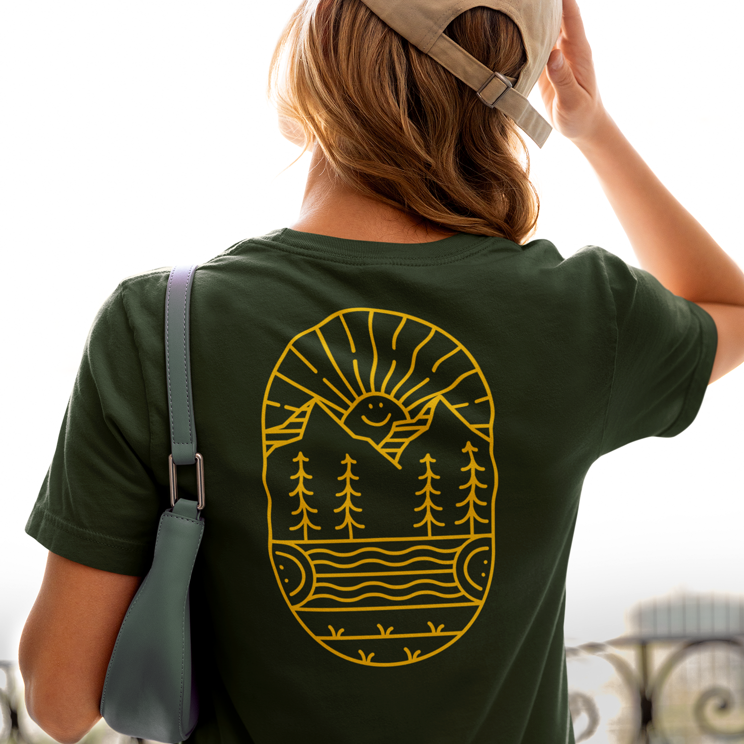 Back of a person wearing a dark green T-shirt with a yellow graphic design of a smiling sun over mountains and trees, and a green shoulder bag, with a beige cap.