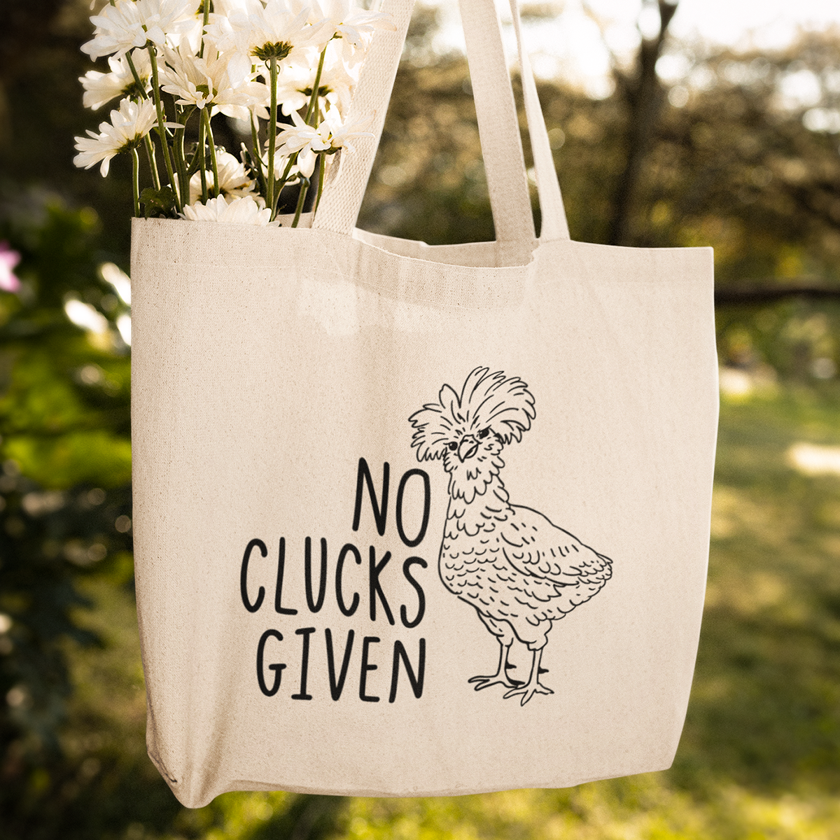 A beige tote bag with a black illustration of a chicken and the words 'NO CLUCKS GIVEN' printed on it, hanging outdoors with white flowers visible inside the bag.
