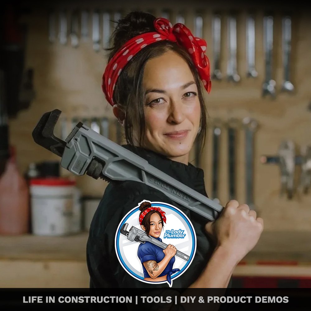 Woman with a red polka dot bandana, holding a large adjustable wrench on her shoulder, standing in a woodworking shop with tools hanging on the wall.