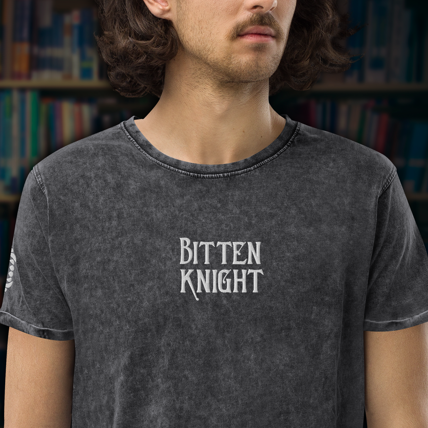 A person wearing a dark gray T-shirt with the words "Bitten Knight" printed on the front, standing in front of bookshelves in a library.