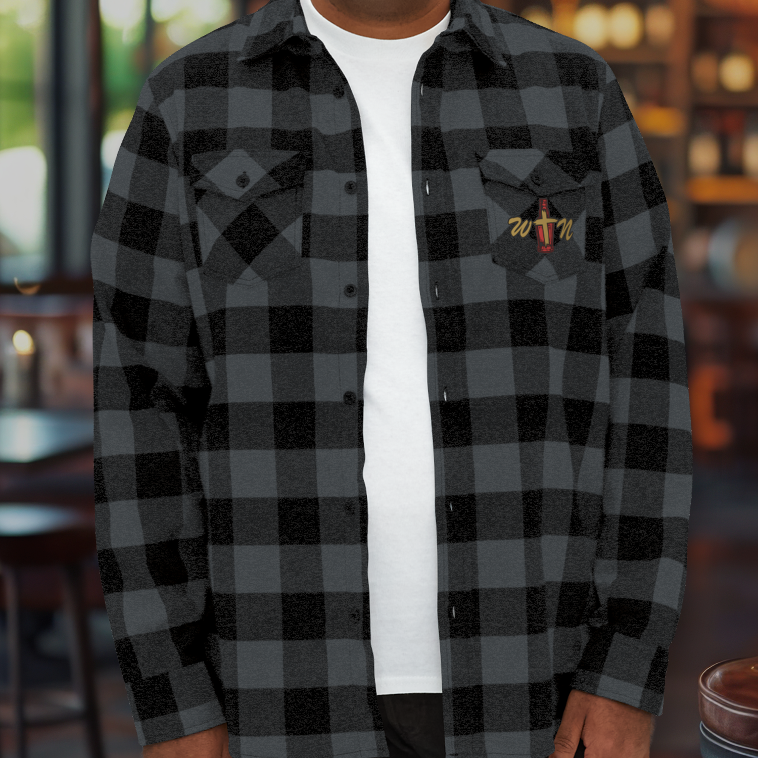 Man wearing a black and gray checkered flannel shirt with a logo on the chest, inside a warmly lit restaurant.
