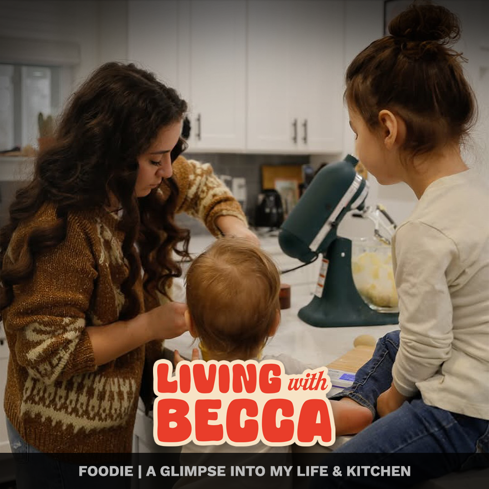 Three women in a kitchen with a small child, one woman with black curly hair wearing a patterned sweater is leaning over a countertop, another with brown hair in a bun, wearing a white shirt, is sitting on the counter, and a toddler with light brown 