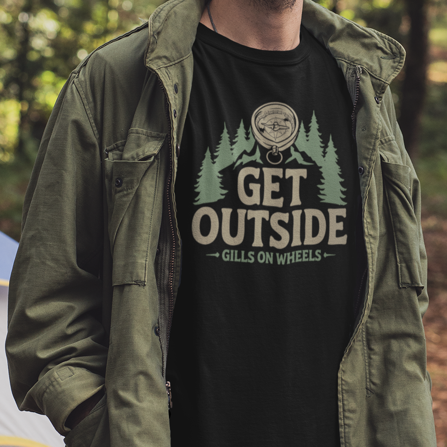Person wearing a green jacket and a black t-shirt with the phrase "Get Outside Gills on Wheels" and a graphic of trees, mountains, and a compass, outdoors in a forest.