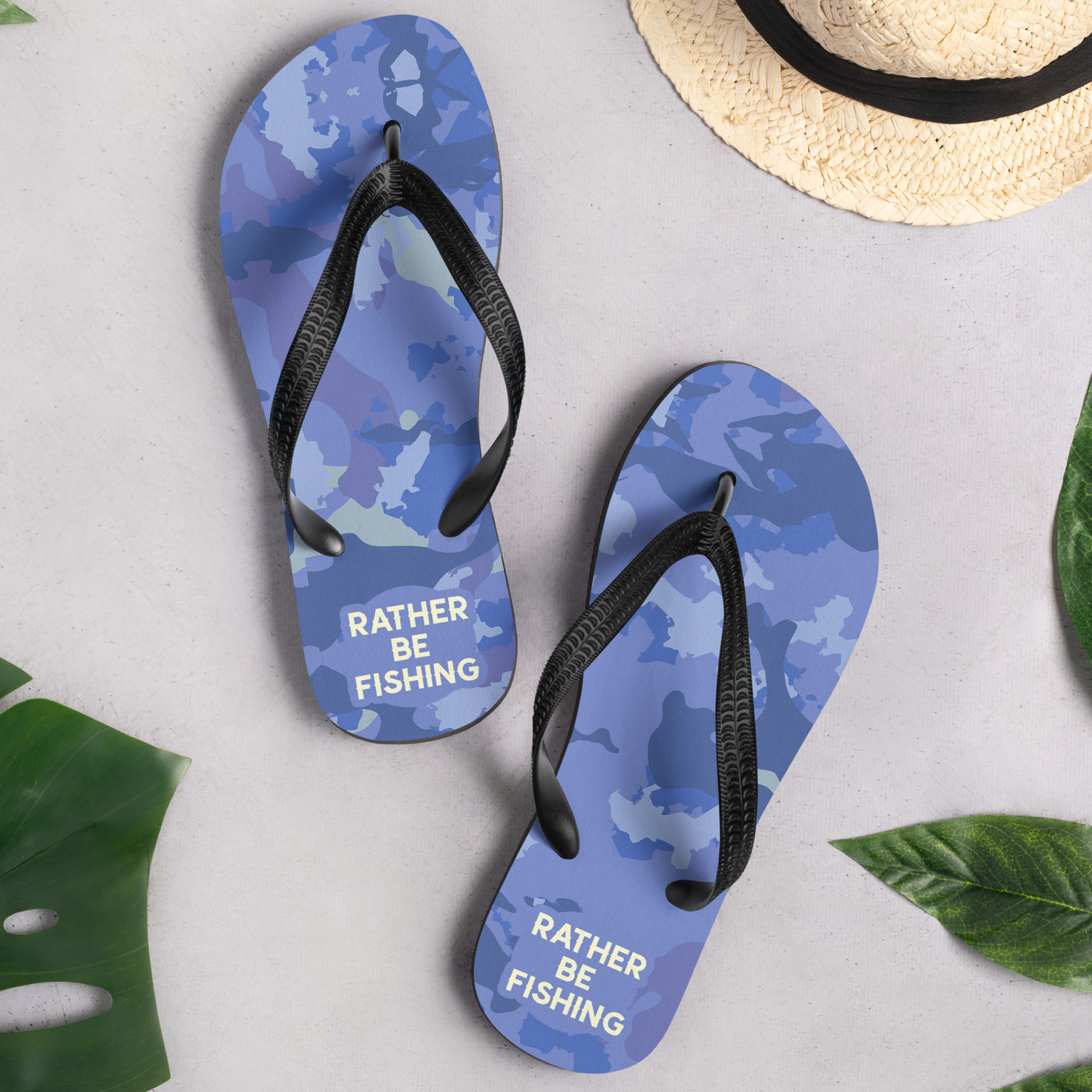Blue flip-flops with camouflage pattern and black straps, placed on a white surface next to a straw hat with a black band and green leaves.
