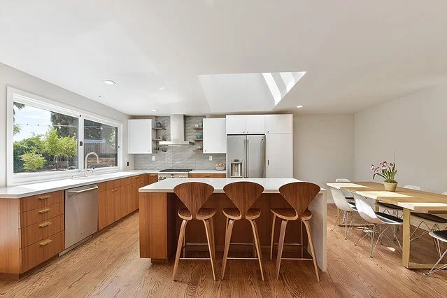 Modern kitchen with wooden island, four barstools, white cabinets, stainless steel appliances, and a dining table with six chairs.