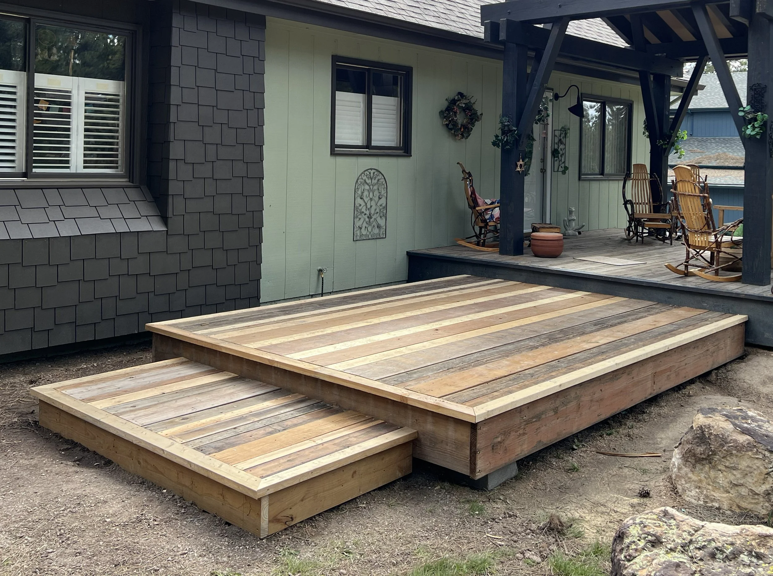 A newly built wooden deck in the backyard of a house, with a raised platform and a smaller step-down section, situated in front of a screened porch with outdoor chairs and decorative items.