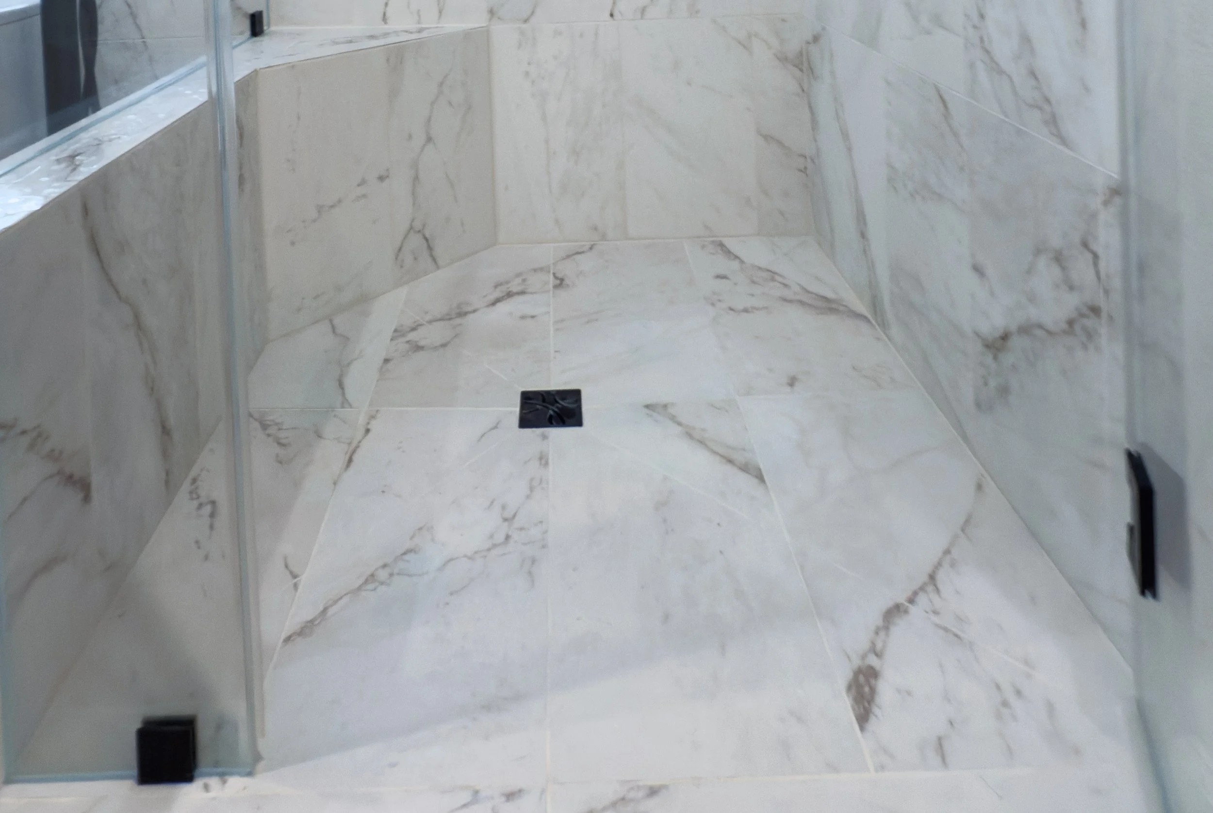 Empty walk-in shower with marble tiles on the walls and floor, a square drain in the center, and glass door on the side.