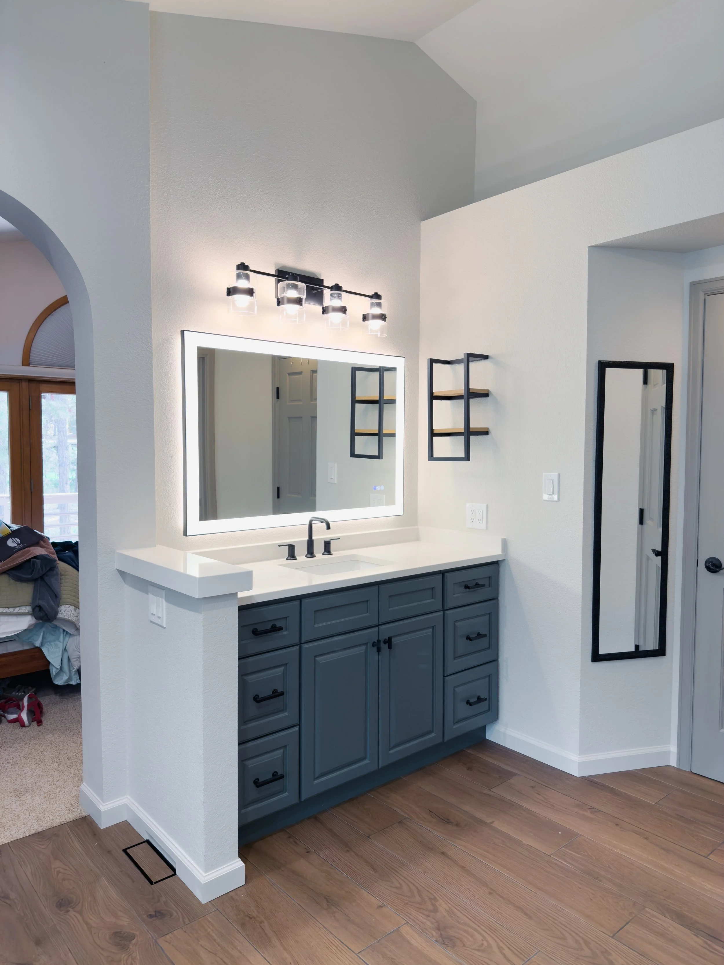 Modern bathroom vanity with blue cabinets, white countertop, and built-in mirror with LED lighting. Black light fixture and wall shelves are visible, with a full-length mirror on the wall."