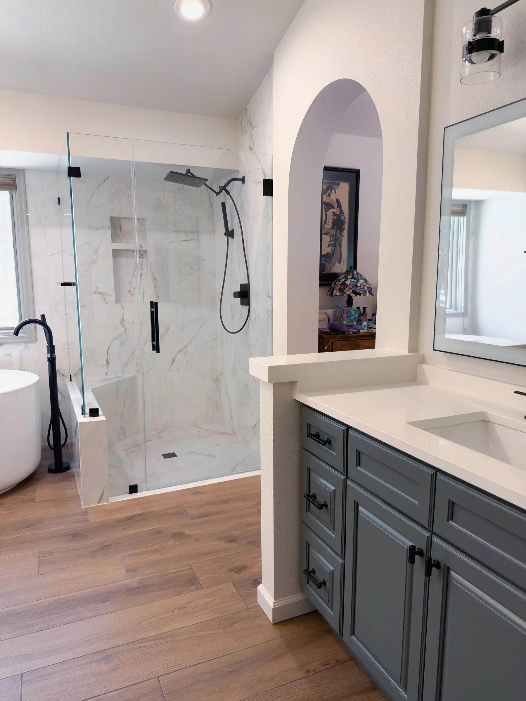 Modern bathroom with a walk-in shower with glass doors, a white vanity with gray cabinets, a large mirror, and wooden flooring. A small arched opening leads to a bedroom with a framed picture and a table lamp.